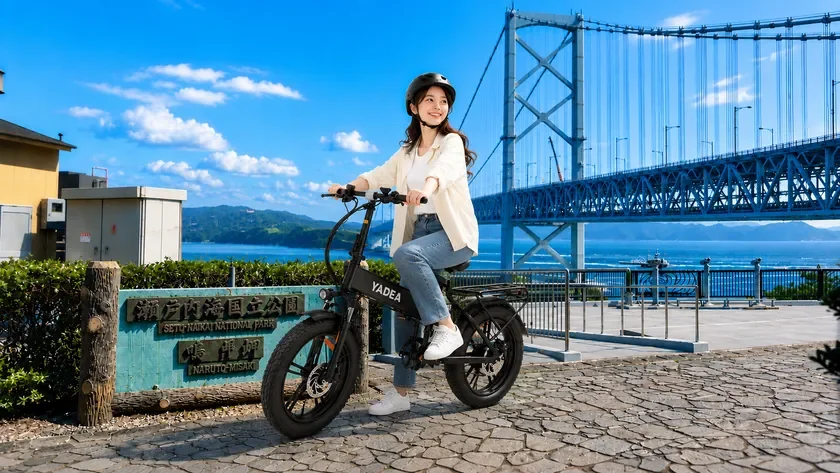 青空の下、女性が黒い電動自転車YADEAに乗って笑顔でポーズをとっています。背景には瀬戸内海と大鳴門橋、そして「瀬戸内海国立公園 鳴門岬」の看板が見える景色の良い場所です。